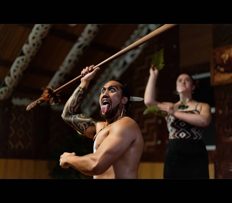 Maori warrior performing a traditional haka with spear and fierce expression.