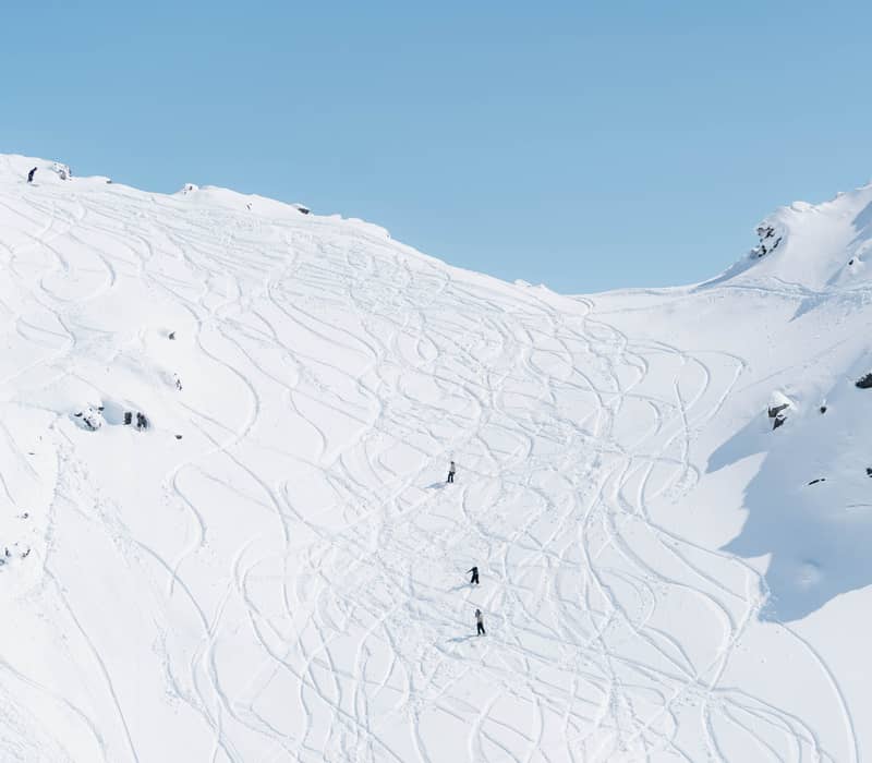 A wide view of a steep, snow-covered mountain bowl at The Remarkables, crisscrossed with dozens of ski and snowboard tracks, with small figures of people at the top of the ridge and descending the slope.