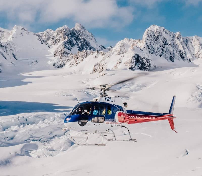 Helicopter flying over a snow-covered glacier with rugged mountain peaks in the background.