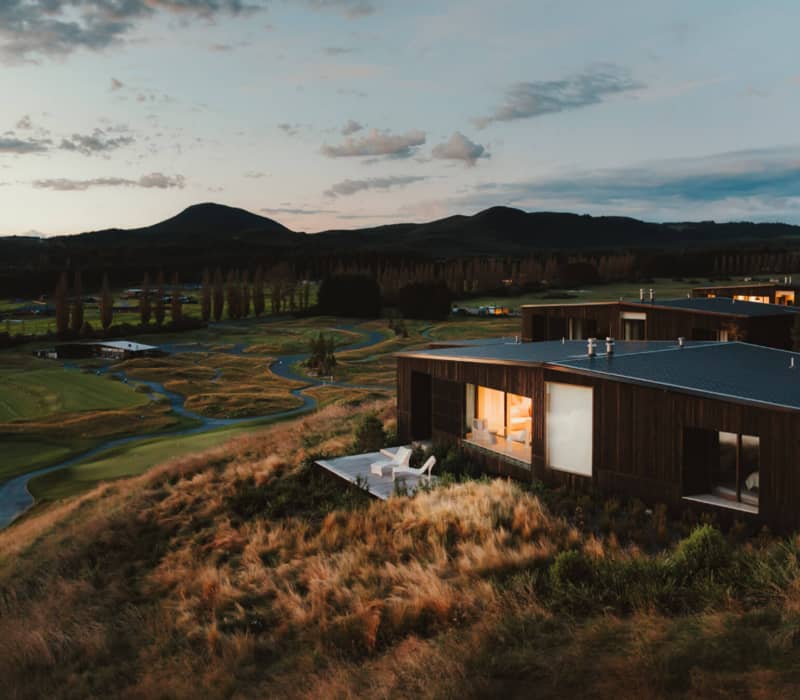 Modern lodge on a grassy hill beside a golf course with mountain views at sunset.