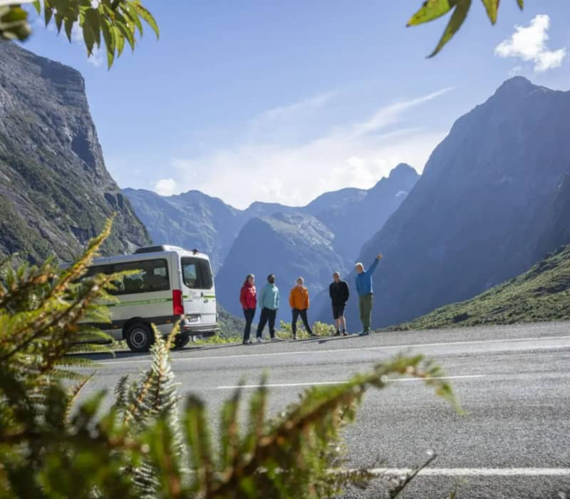 A Trips & Tramps tour guide points toward towering mountain peaks for a small group of hikers standing next to their tour van.