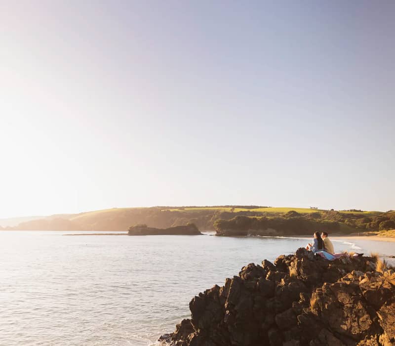 Couple sitting on coastal rocks overlooking the ocean at sunset.