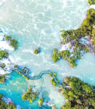 An aerial bird's-eye view of a winding wooden boardwalk crossing through vibrant turquoise and pale green geothermal pools surrounded by dense green native bush.