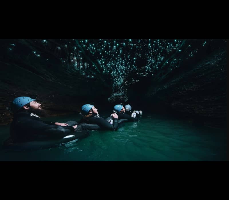 A line of adventurers in wetsuits and blue helmets float on black inner tubes down a subterranean river, gazing up at a dense canopy of glowing blue glow worms on the cave ceiling.