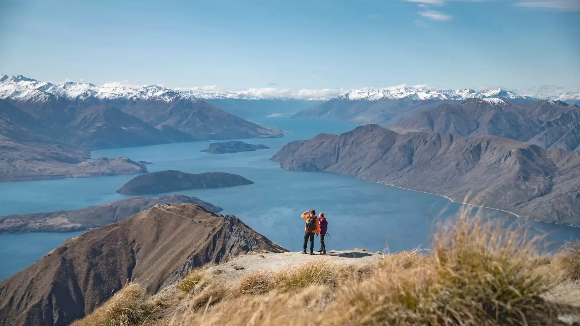 Two hikers on Roy's Peak with a wide view of Lake Wanaka and snowy mountains in the background.
