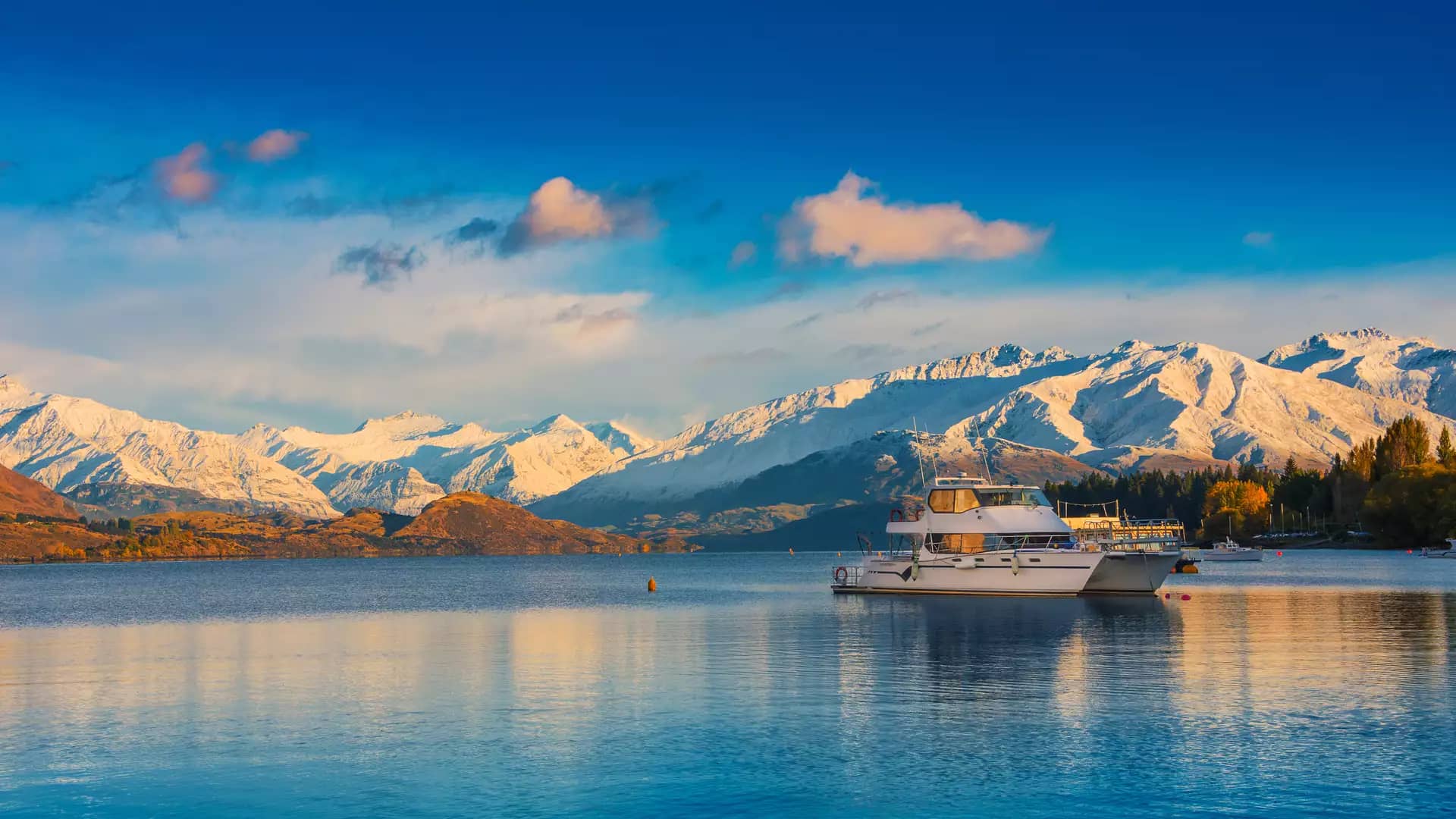Boat on calm Lake Wanaka with snow-covered mountains and autumn trees at sunset.