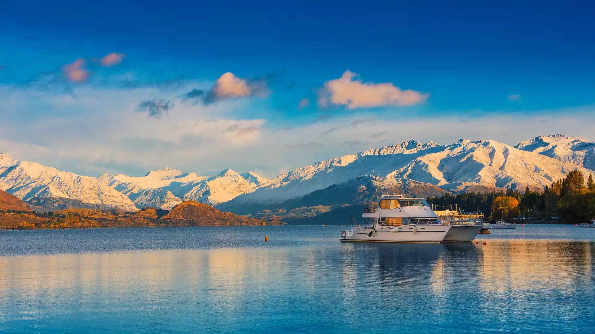 Boat on calm Lake Wanaka with snow-covered mountains and autumn trees at sunset.