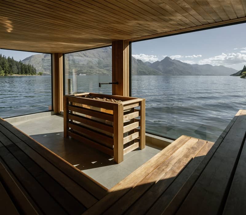 The interior of a modern wood-paneled sauna featuring tiered seating and a central heater, with a large glass window offering a panoramic view of a calm lake and rugged mountains.