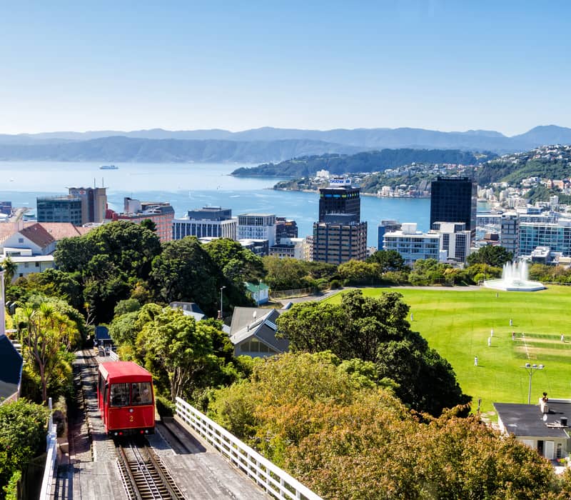 Red cable car moves up hill with city, cricket field and harbor in background under clear sky.