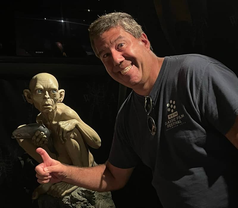 A man in a navy t-shirt smiles and gives a thumbs-up next to a highly detailed, life-sized statue of Gollum holding a fish, set against a dark background at the Wētā Cave in Wellington.