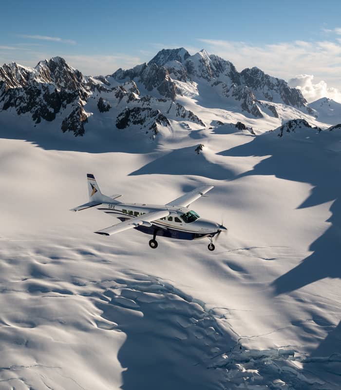 Small plane flying over snowy glacier and rugged mountain peaks under clear sky
