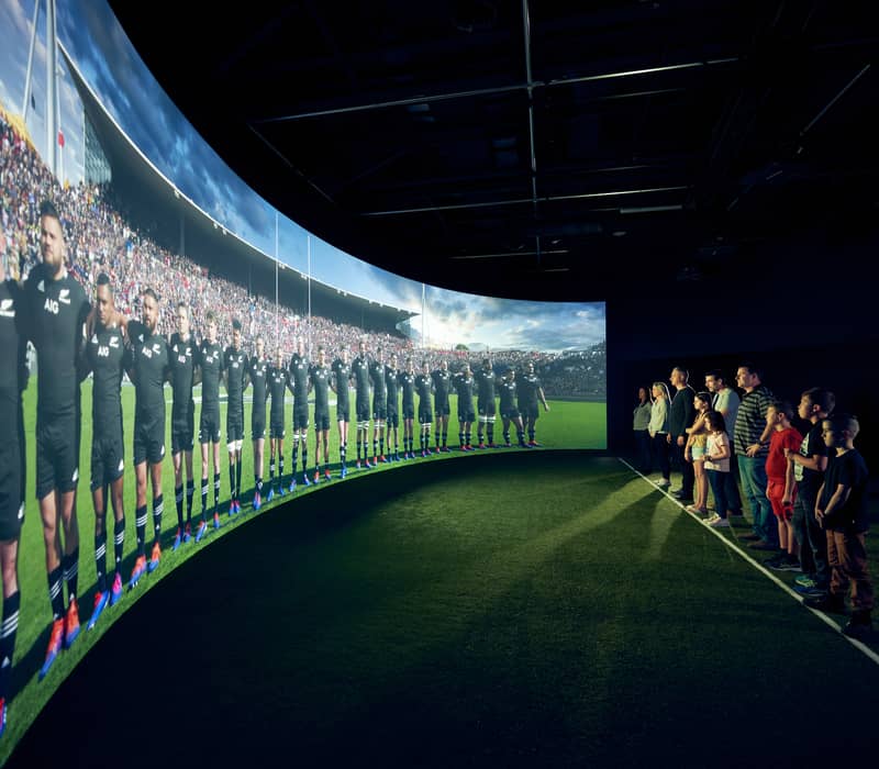 People watching a large screen of rugby players lined up on a field in a stadium setting.
