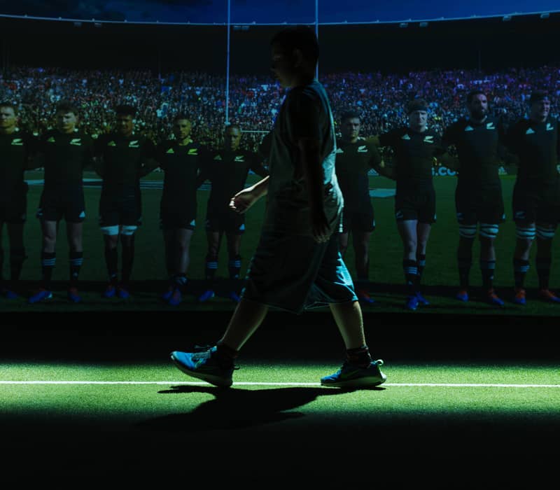 Child walking on green field with projection of rugby players lined up in stadium