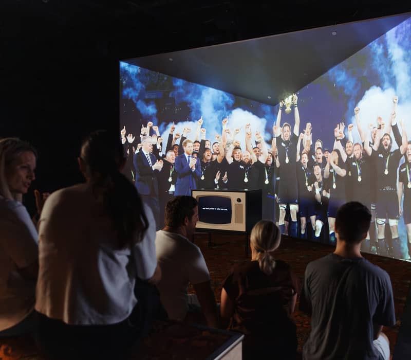 Group of people seated watching a large projected image of rugby players celebrating on a dark indoor floor.