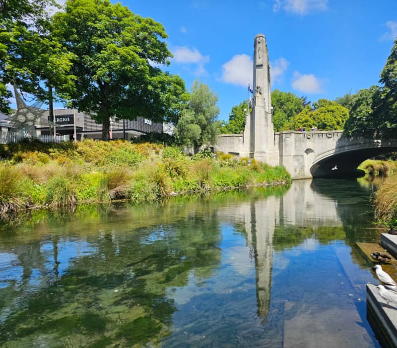 Stone bridge and monument over clear river with trees and birds under a blue sky.