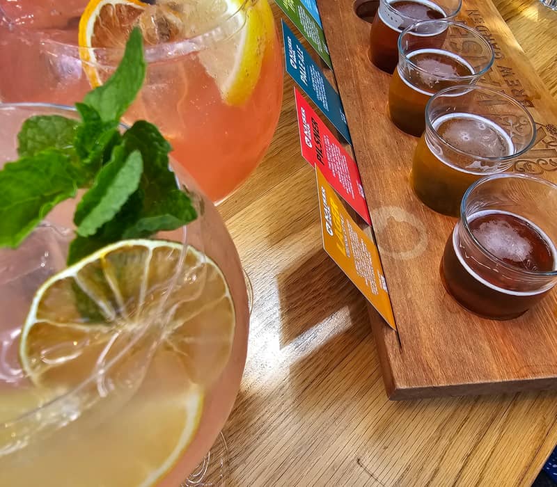 Two cocktails with lemon and mint garnishes and a wooden board holding five small beer glasses with a tasting menu.