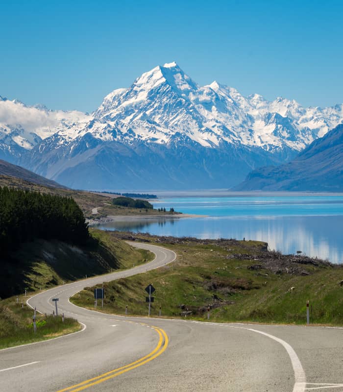 Curved road leading towards snow-covered Aoraki/Mt. Cook mountains by a reflective lake.