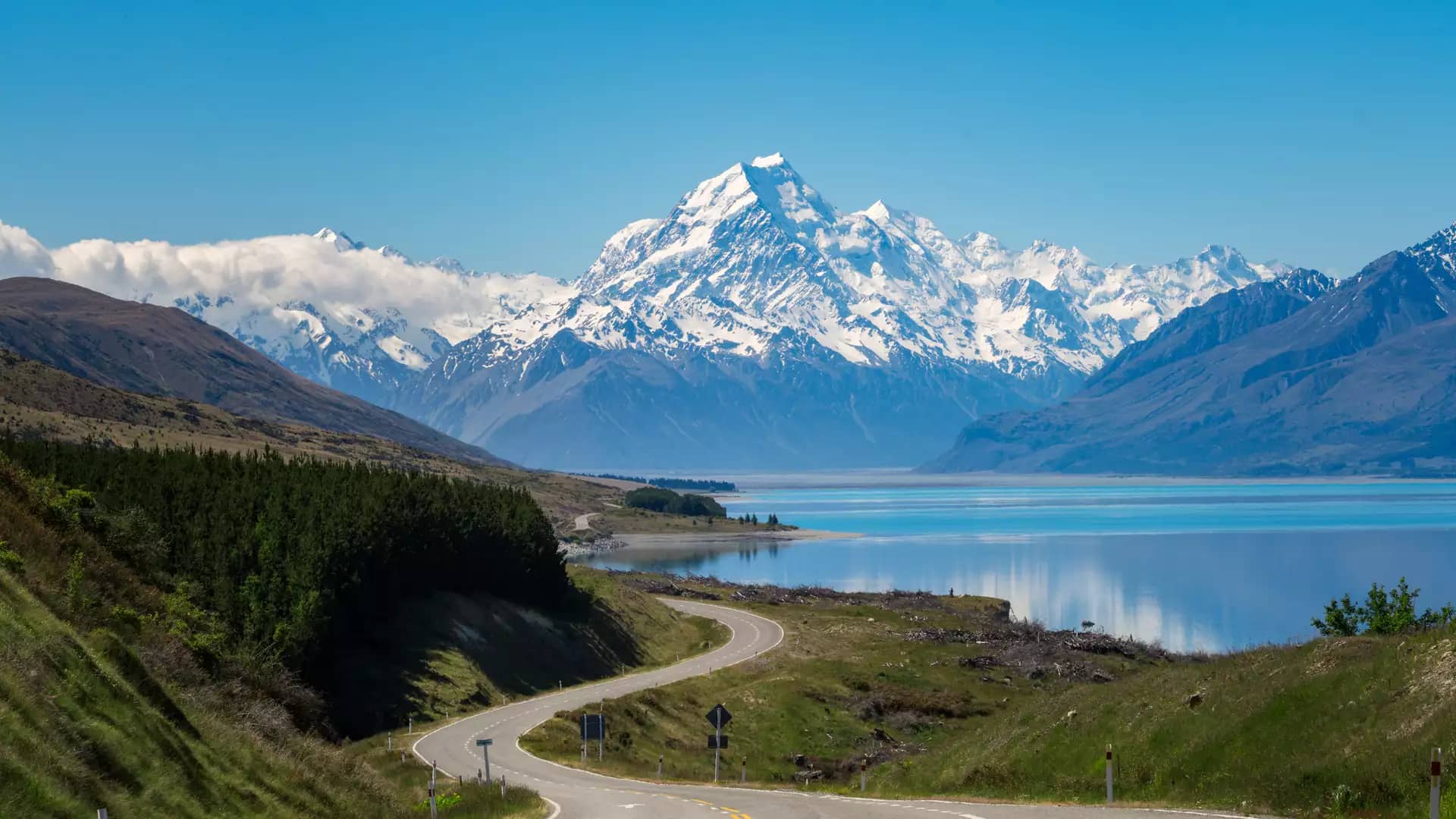 Curved road leading towards snow-covered Aoraki/Mt. Cook mountains by a reflective lake.