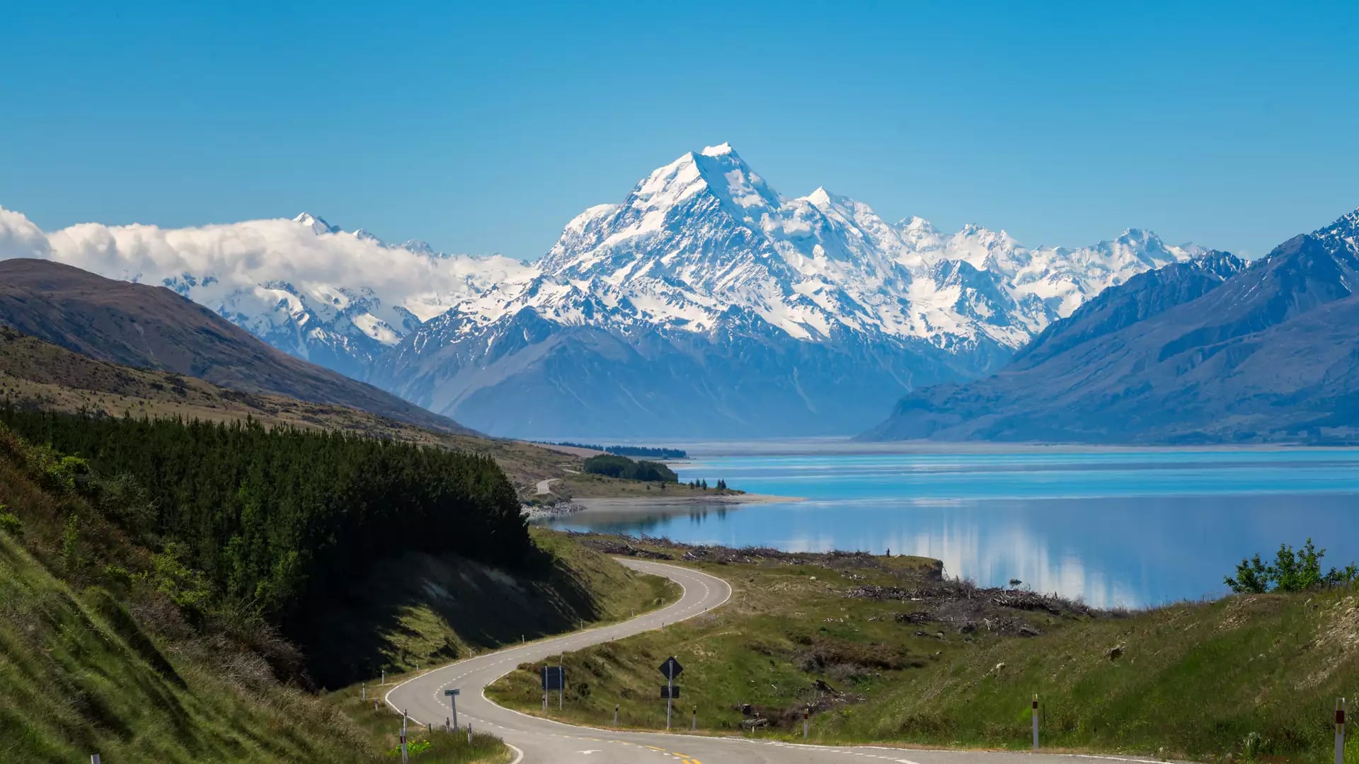 Curved road leading towards snow-covered Aoraki/Mt. Cook mountains by a reflective lake.