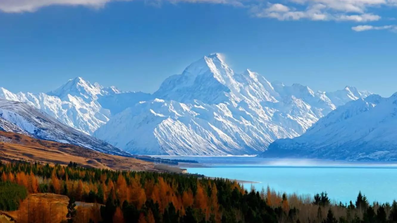 Snow-covered mountain peaks above a turquoise lake with mixed forest in foreground under a blue sky.