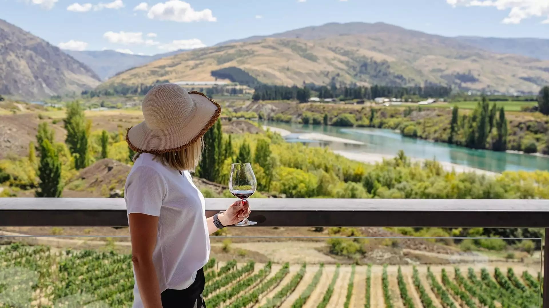 Woman with sun hat holding a glass of wine overlooking vineyard, river, and mountains.