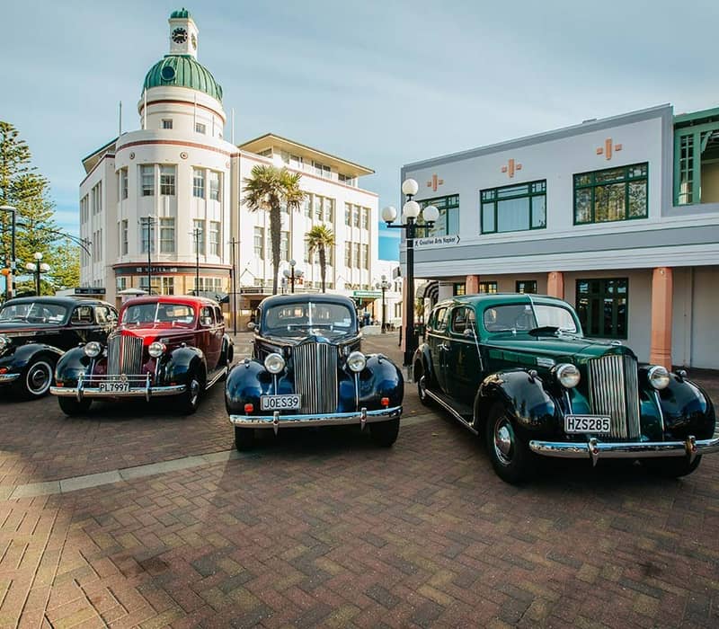 Four classic vintage cars parked outside Art Deco buildings with a clock tower and palm trees on a sunny day.