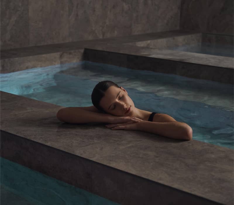 Woman resting her head on her arms at the edge of an indoor marble bathhouse pool.