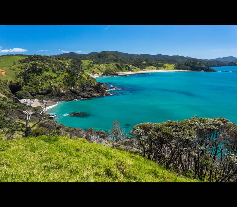 Turquoise bay surrounded by green hills and trees with a clear blue sky.