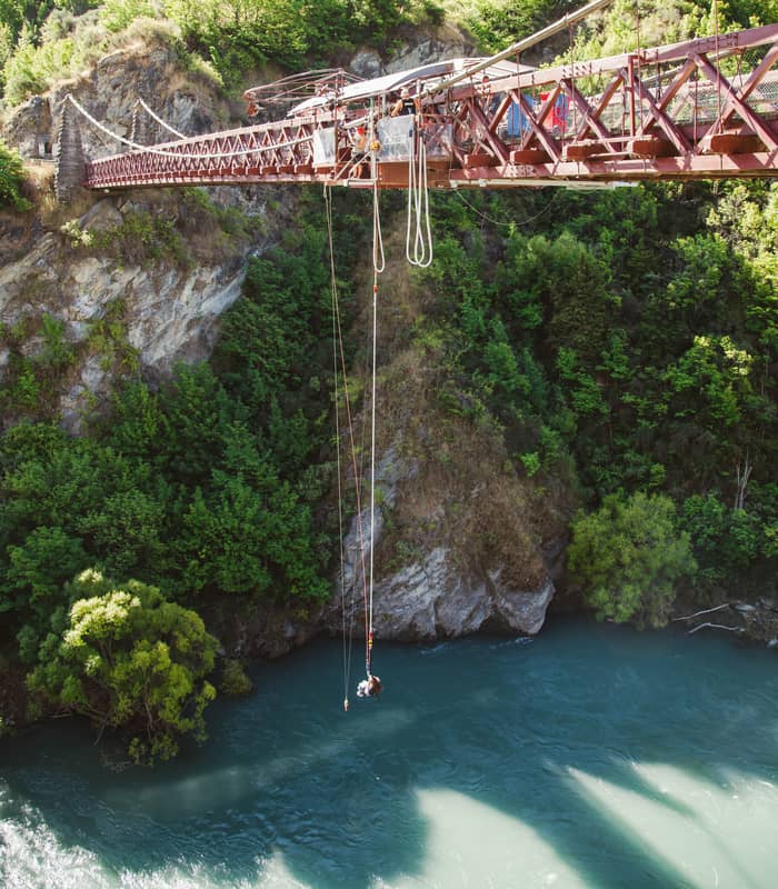 Person bungy jumping from a bridge over a river with mountainous forest background.
