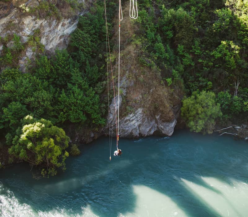 Person bungy jumping from a bridge over a river with mountainous forest background.