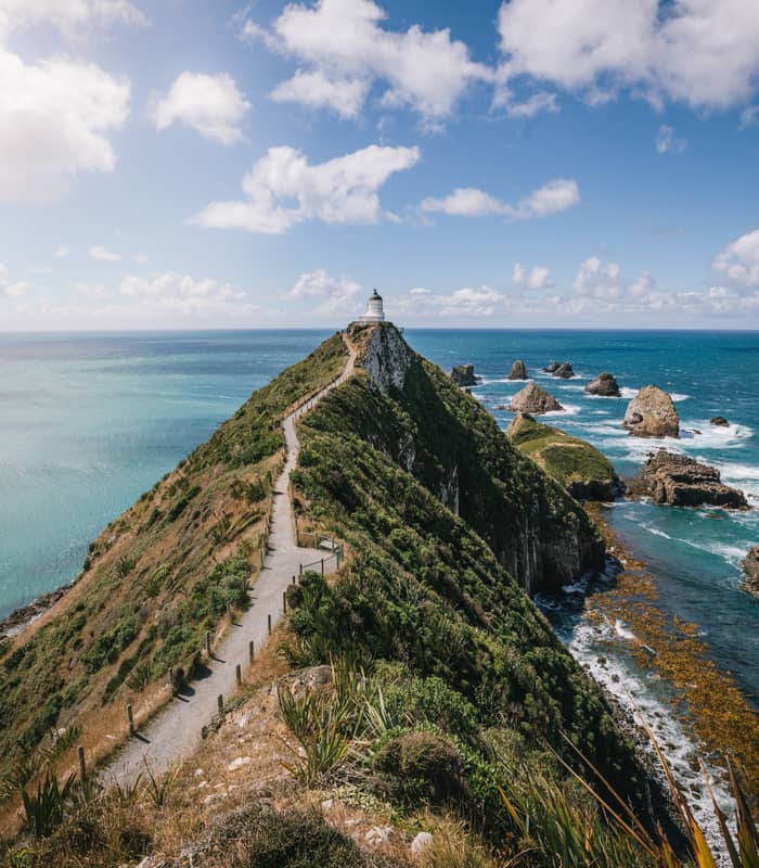 Lighthouse atop a coastal cliff with a walking path and ocean view under a partly cloudy sky.
