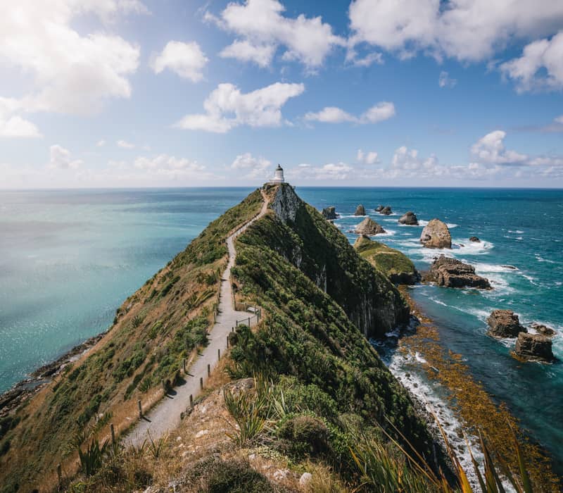 Lighthouse atop a coastal cliff with a walking path and ocean view under a partly cloudy sky.