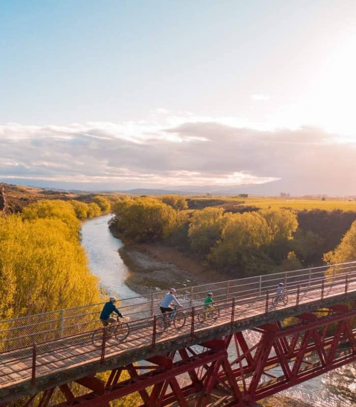 Family cycling over Manuherikia Bridge with river and trees at sunset