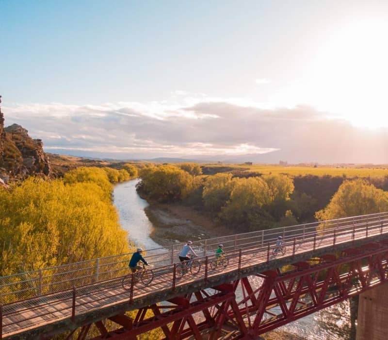 Family cycling over Manuherikia Bridge with river and trees at sunset