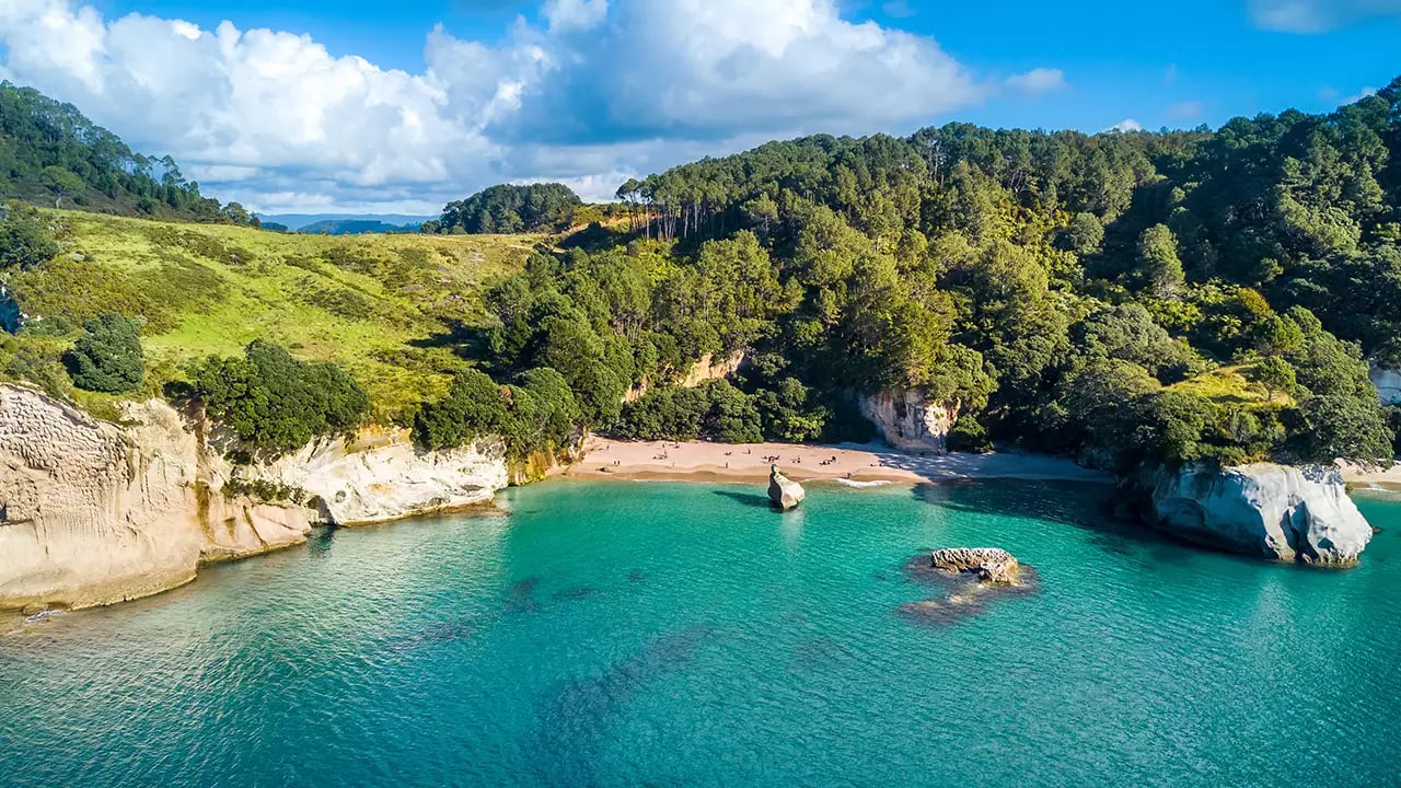 Aerial view of a small beach with turquoise water, cliffs, and forest under a cloudy sky.