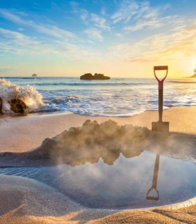 Shovel next to a steaming hot spring pool on a sandy beach with waves and sunset in the background.