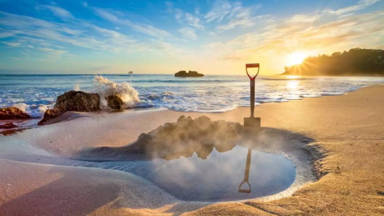 Shovel next to a steaming hot spring pool on a sandy beach with waves and sunset in the background.