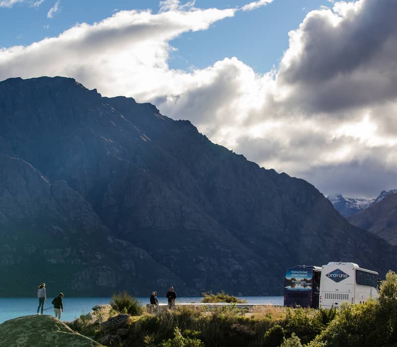 People near Lake Hawea with mountains, cloudy sky, and tour buses parked nearby.