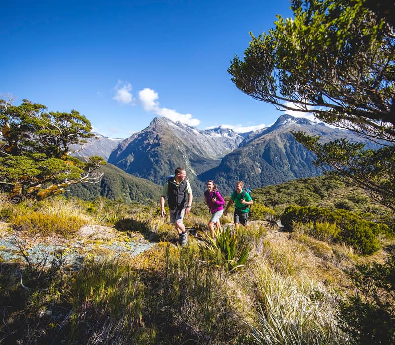 Three hikers walking on a trail with mountainous landscape and clear sky.
