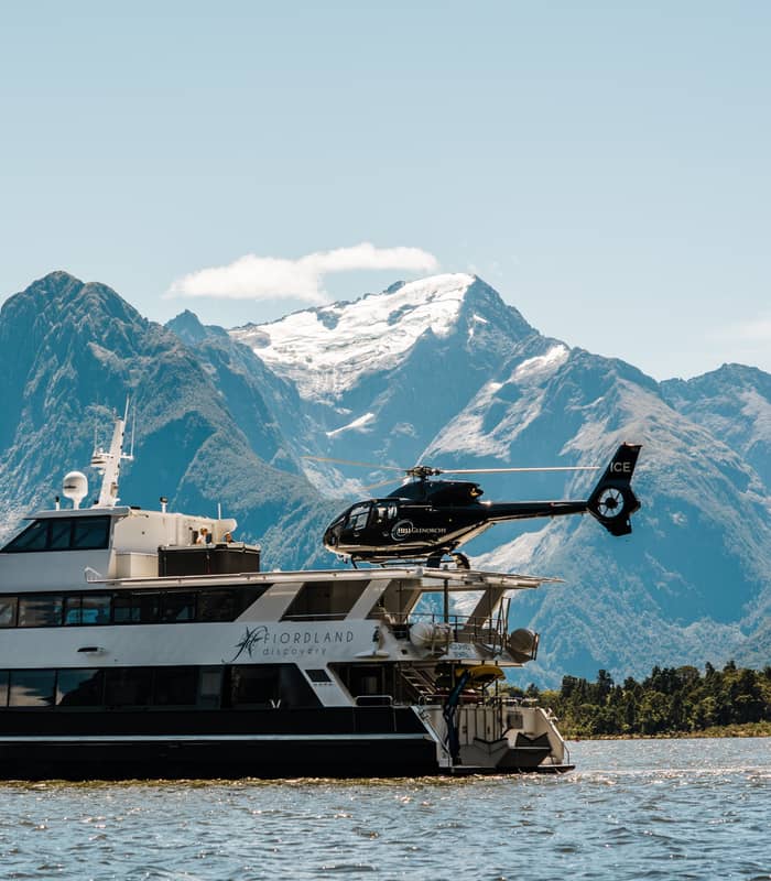 Luxury boat with helicopter on deck on water with tall mountains and blue sky background.