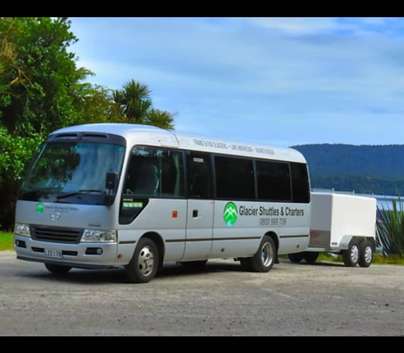 White Glacier Shuttles & Charters bus with trailer parked by a lake with trees and hills in background.