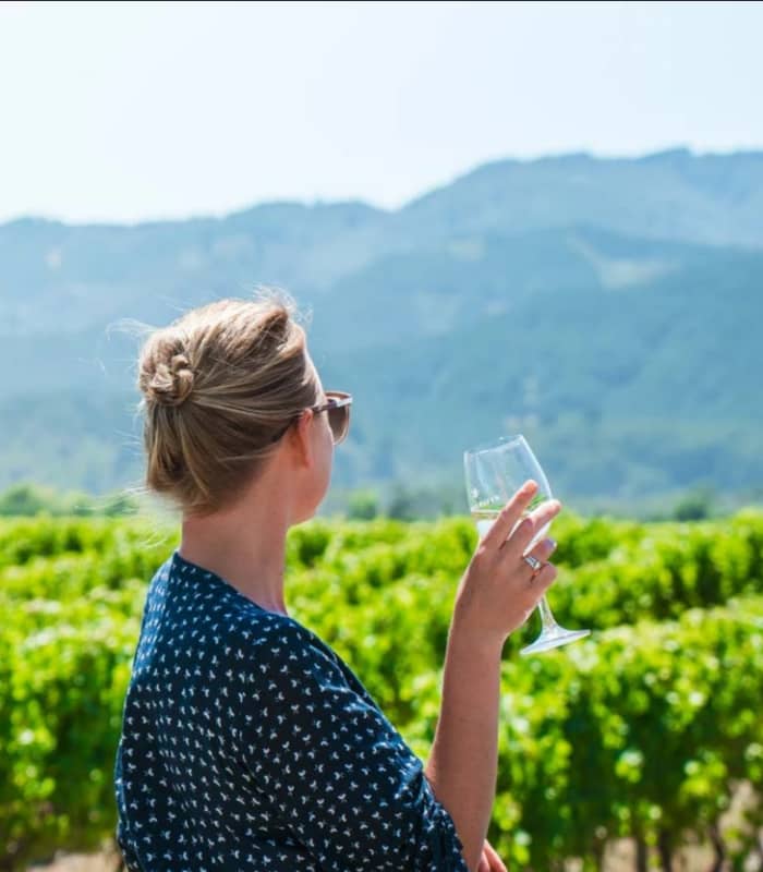 Woman holding a glass of white wine standing in a green vineyard with mountains in the background.