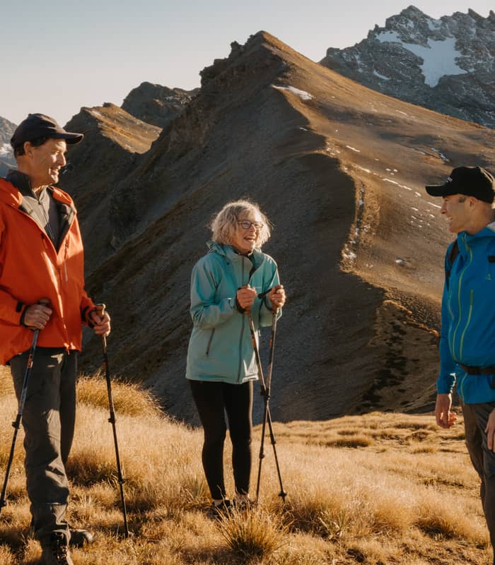 Three hikers standing on a mountain trail with trekking poles, talking and enjoying the sunny weather.