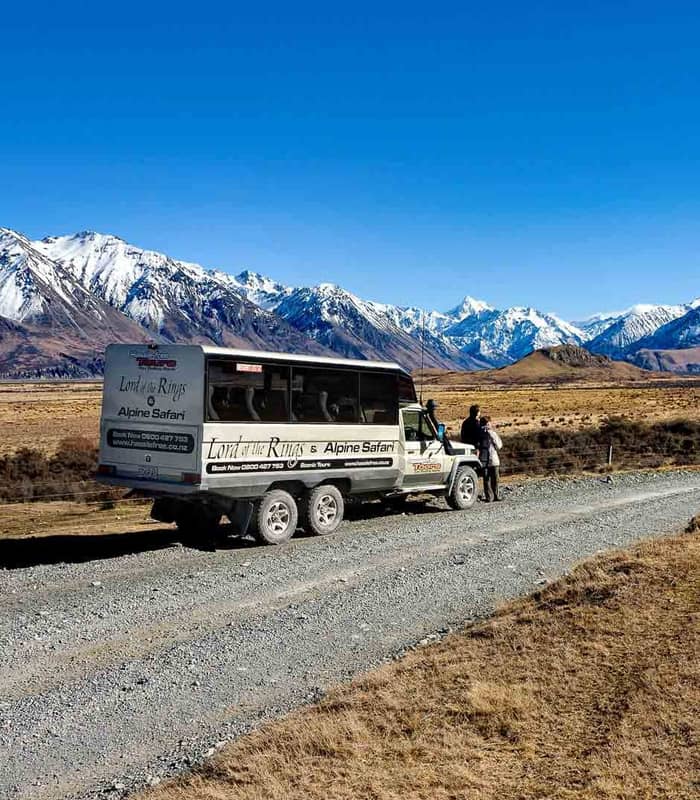 4x4 tour bus parked on a dirt road with people beside it and snow-covered mountains in the background under a clear sky.