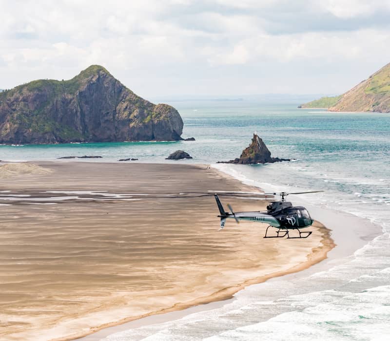 Helicopter flying close to a sandy beach with green islands and ocean waves under a cloudy sky.