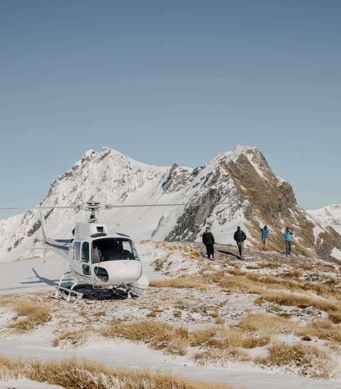 People hiking near a helicopter on a snowy mountain ridge with clear blue sky.