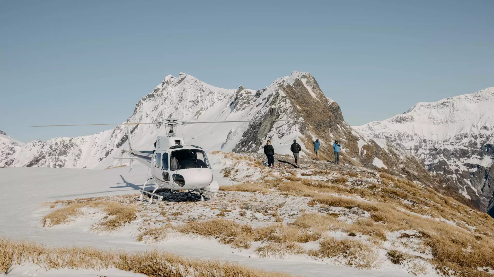 People hiking near a helicopter on a snowy mountain ridge with clear blue sky.