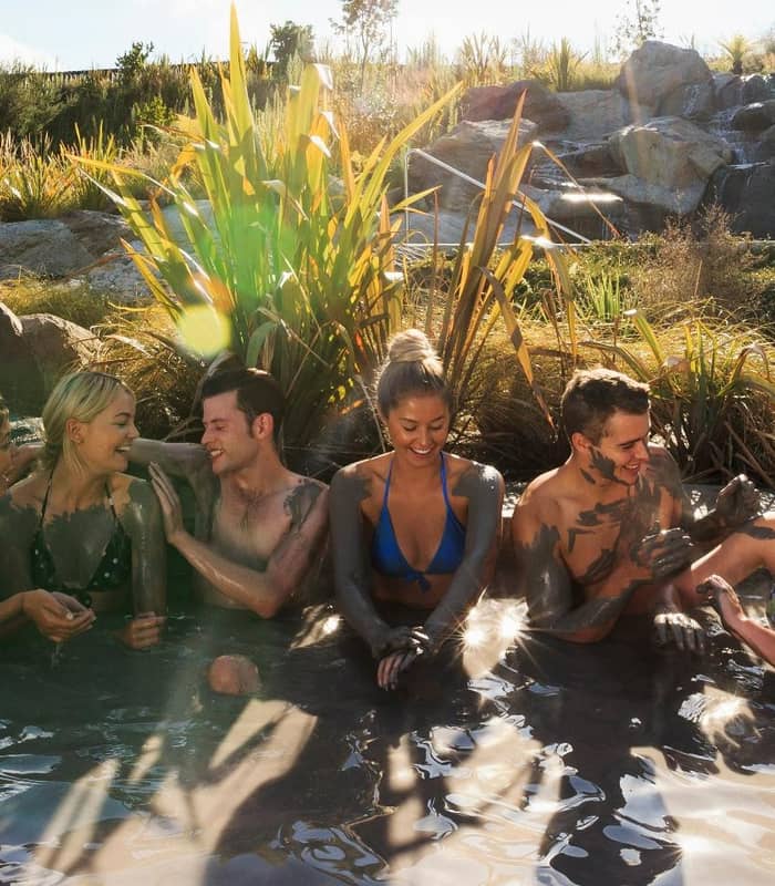 Six people in swimwear covered in mud enjoying an outdoor mud bath with plants and rocks around.