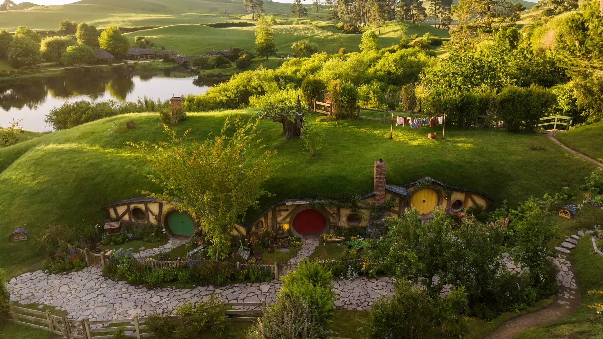 Hobbiton movie set with hobbit holes, gardens, and green hills beside a calm lake at sunset.