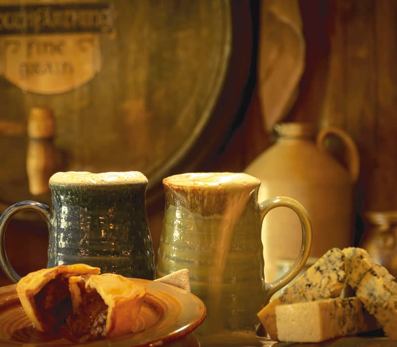 Two mugs of foamy beer with a meat pie and blue cheese on a wooden table.